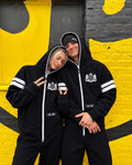 Couple in 69 black jumpsuits by a yellow wall | THE ONE