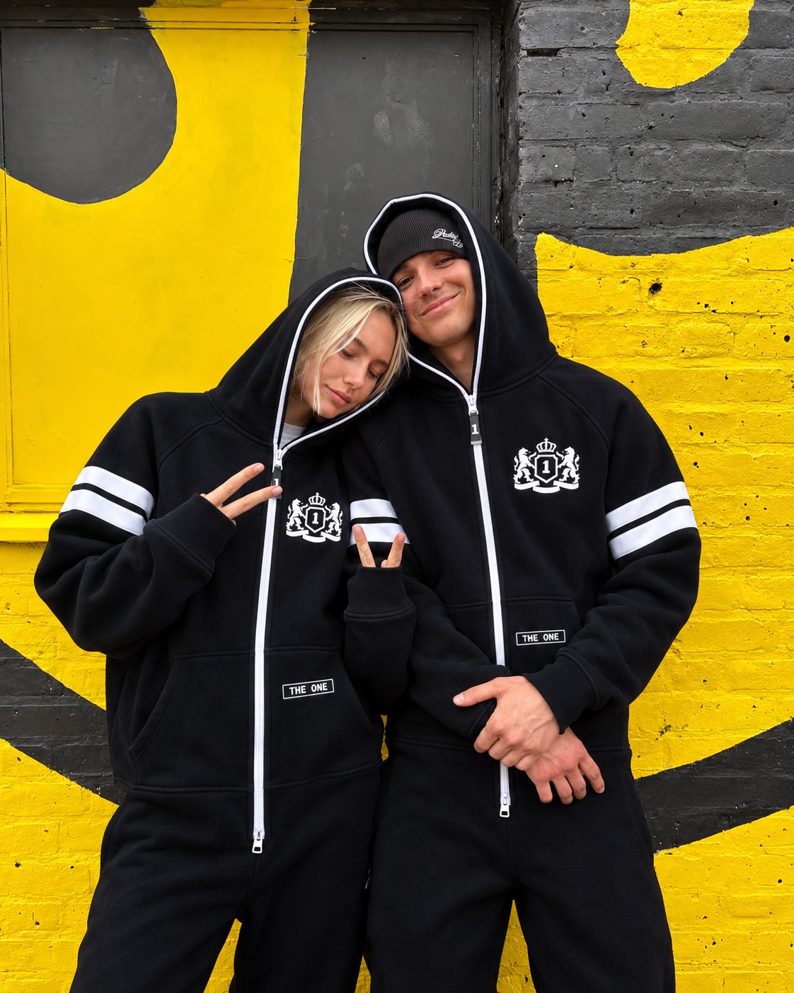 Couple in 69 black jumpsuits by a yellow wall | THE ONE